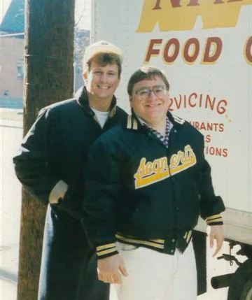 Two men in 1980s casual wear standing together, one wearing an Athletics jacket, representing classic foodservice wholesale distribution era.